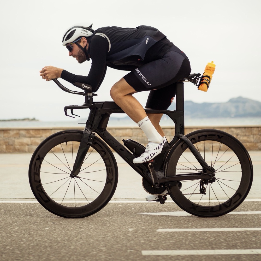 Triathlet in Aeroposition auf dem Zeitfahrrad an der Küste, dahinter Meer und Berge
