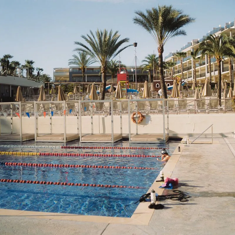 Sportpool unter Palmen: Ein Schwimmer macht Pause am Beckenrand, daneben Trinkflaschen, Pullbuoy und Paddles
