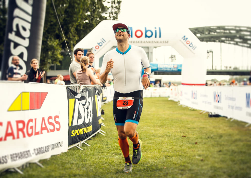 Lächelnder Triathlet im weißen Einteiler beim Zieleinlauf des Cologne 226 Langdistanz-Triathlons in Köln