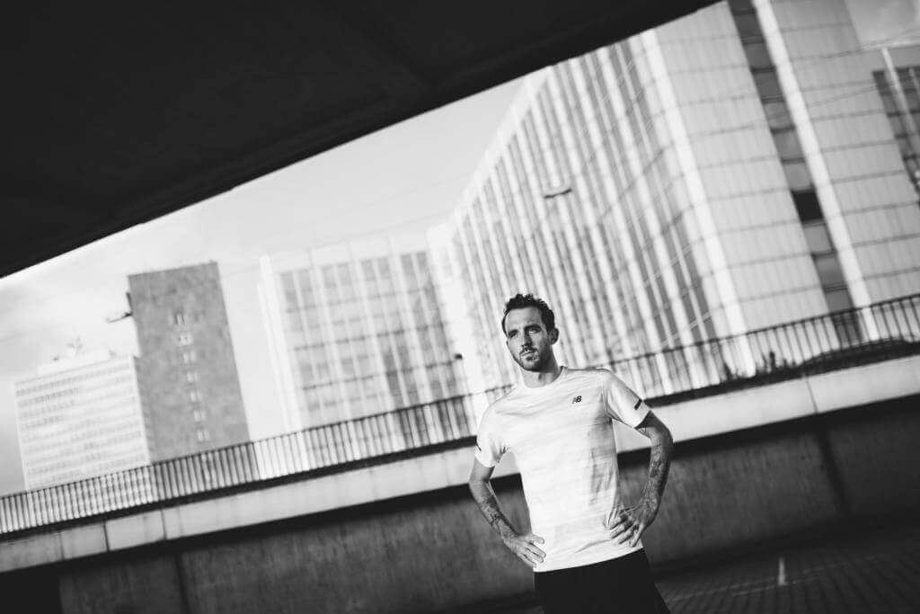Niclas Bock im weißen Laufshirt, die Hände in die Hüften gestützt, vor Hochhäusern unter einer Brücke, Schwarz-Weiß-Foto