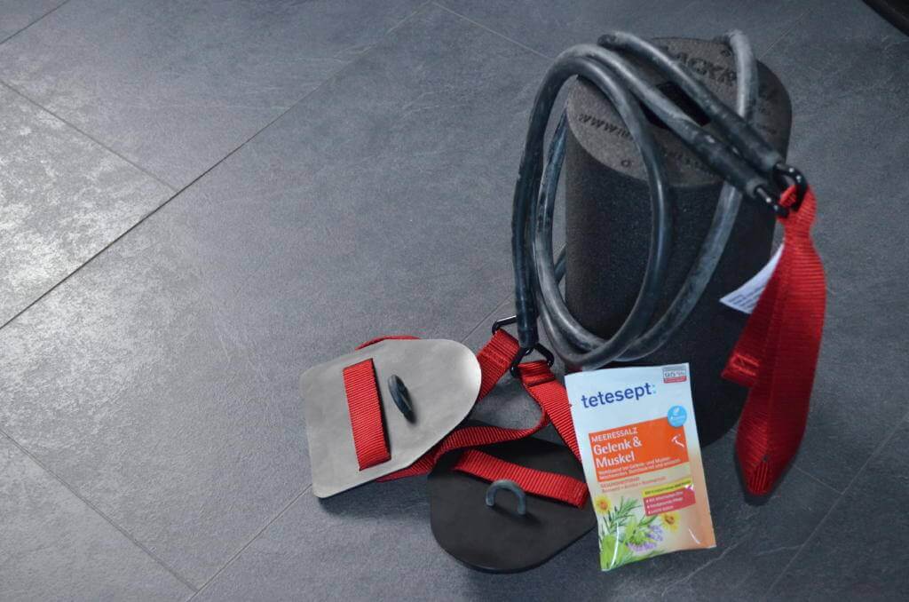Trainingsequipment auf Fliesenboden: Handpaddles, Zugseil mit roten Schlaufen, Faszienrolle und Badesalz von tetesept