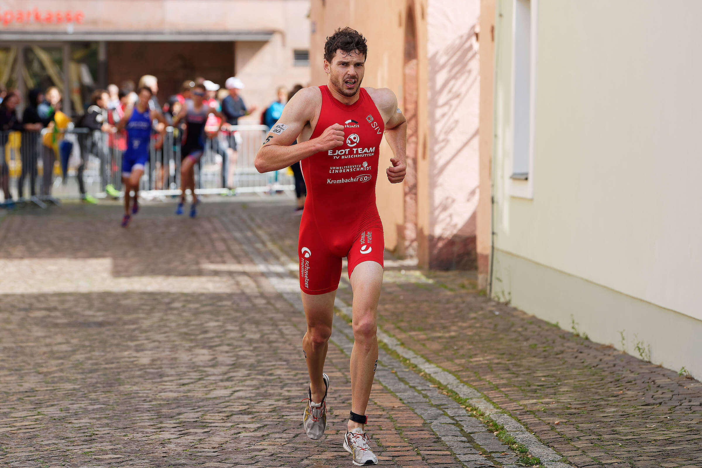 Franz Löschke im roten Einteiler des EJOT Teams auf der Laufstrecke eines Triathlons über Kopfsteinpflaster