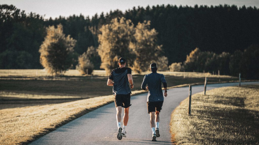 Zwei Läufer von hinten auf einem asphaltierten Weg durch Wiesen und Wald im warmen Abendlicht