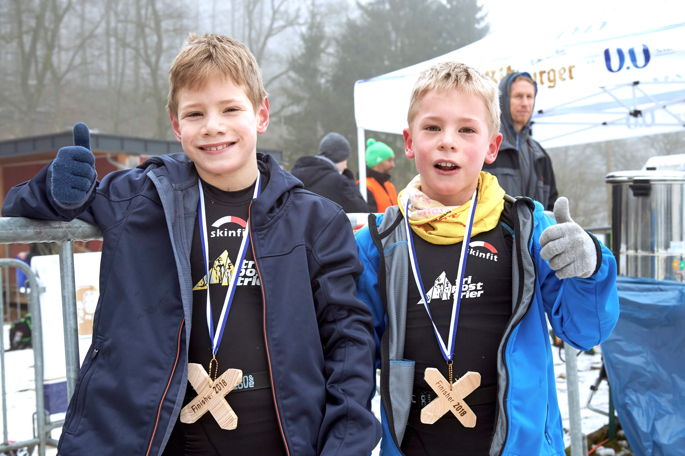 Zwei Kinder mit hölzernen Finisher-Medaillen zeigen bei einem winterlichen Wettkampf den Daumen nach oben