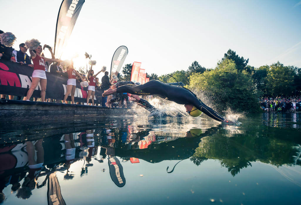 Triathleten springen beim Schwimmstart des Ironman 70.3 St. Pölten 2018 vom Steg in den See, dahinter Cheerleader