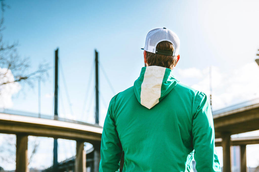 Mann mit weißer Basecap und grüner Jacke von hinten, er blickt auf eine Schrägseilbrücke unter blauem Himmel