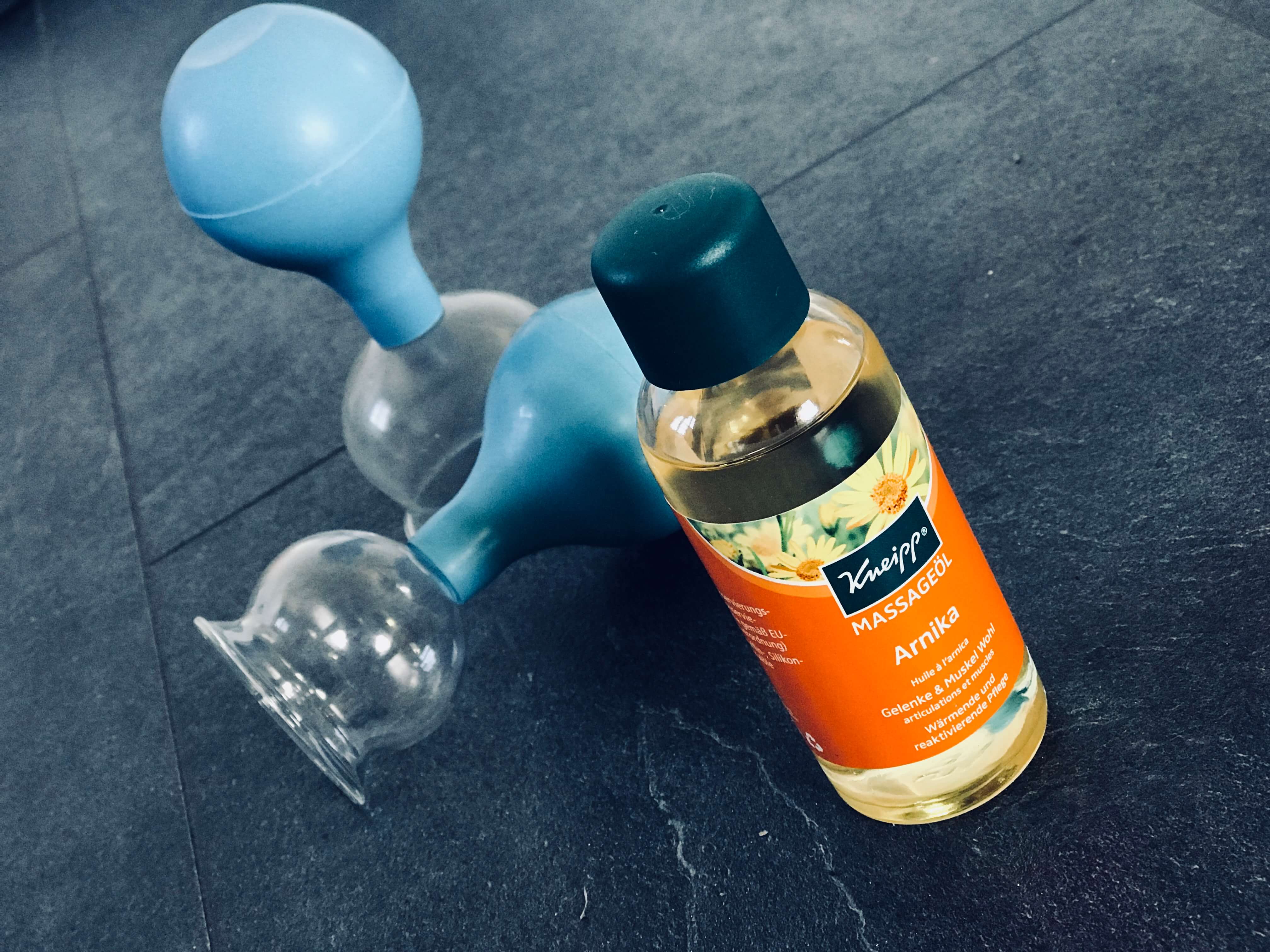 Zwei Schröpfgläser mit blauem Saugball und eine Flasche Kneipp Massageöl Arnika auf dunklem Stein