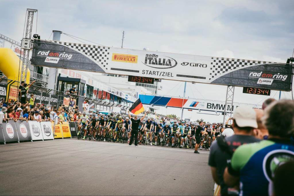Startaufstellung bei Rad am Ring 2018 unter dem Start-Ziel-Banner am Nürburgring, ein Starter schwenkt die Deutschlandfahne