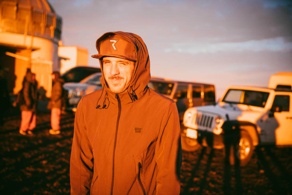 Mann in orangefarbener Kapuzenjacke mit Kappe im goldenen Abendlicht, dahinter Geländewagen und ein Kuppelbau