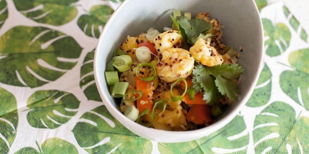 Fruchtige Quinoa-Pfanne mit Garnelen, Ananas, Paprika und Frühlingszwiebeln in weißer Schale auf grünem Blattmuster