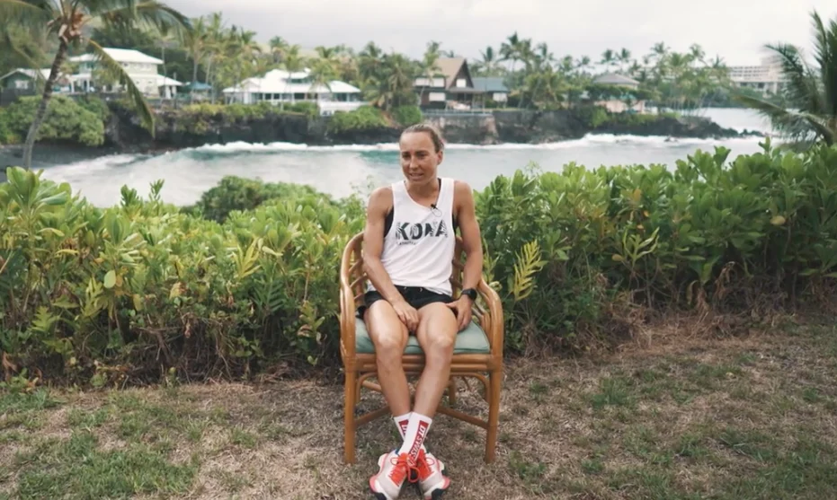 Anne Haug im Kona-Shirt sitzt für ihre Doku auf einem Rattanstuhl vor einer Bucht mit Palmen auf Hawaii