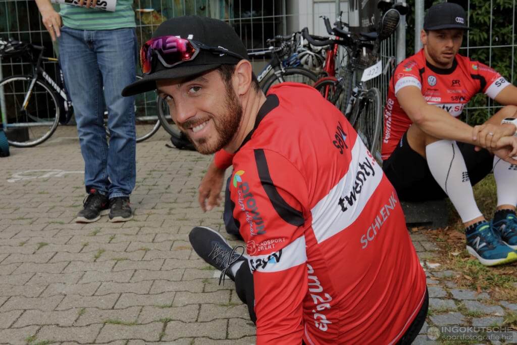 Niclas Bock im roten Teamtrikot sitzt lachend auf dem Pflaster vor Radständern, dahinter ein weiterer Athlet