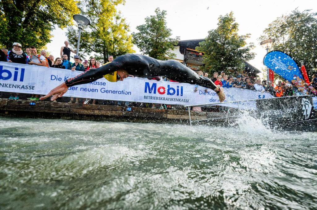 Triathlet im Neoprenanzug springt kopfüber ins Wasser, dahinter Zuschauer und Banner der BKK Mobil Oil