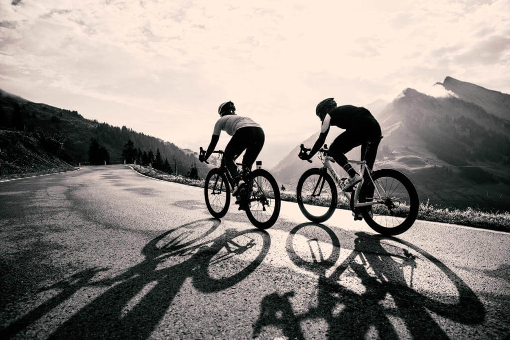 Zwei Rennradfahrer im Gegenlicht auf einer Passstraße am Furkapass, lange Schatten auf dem Asphalt