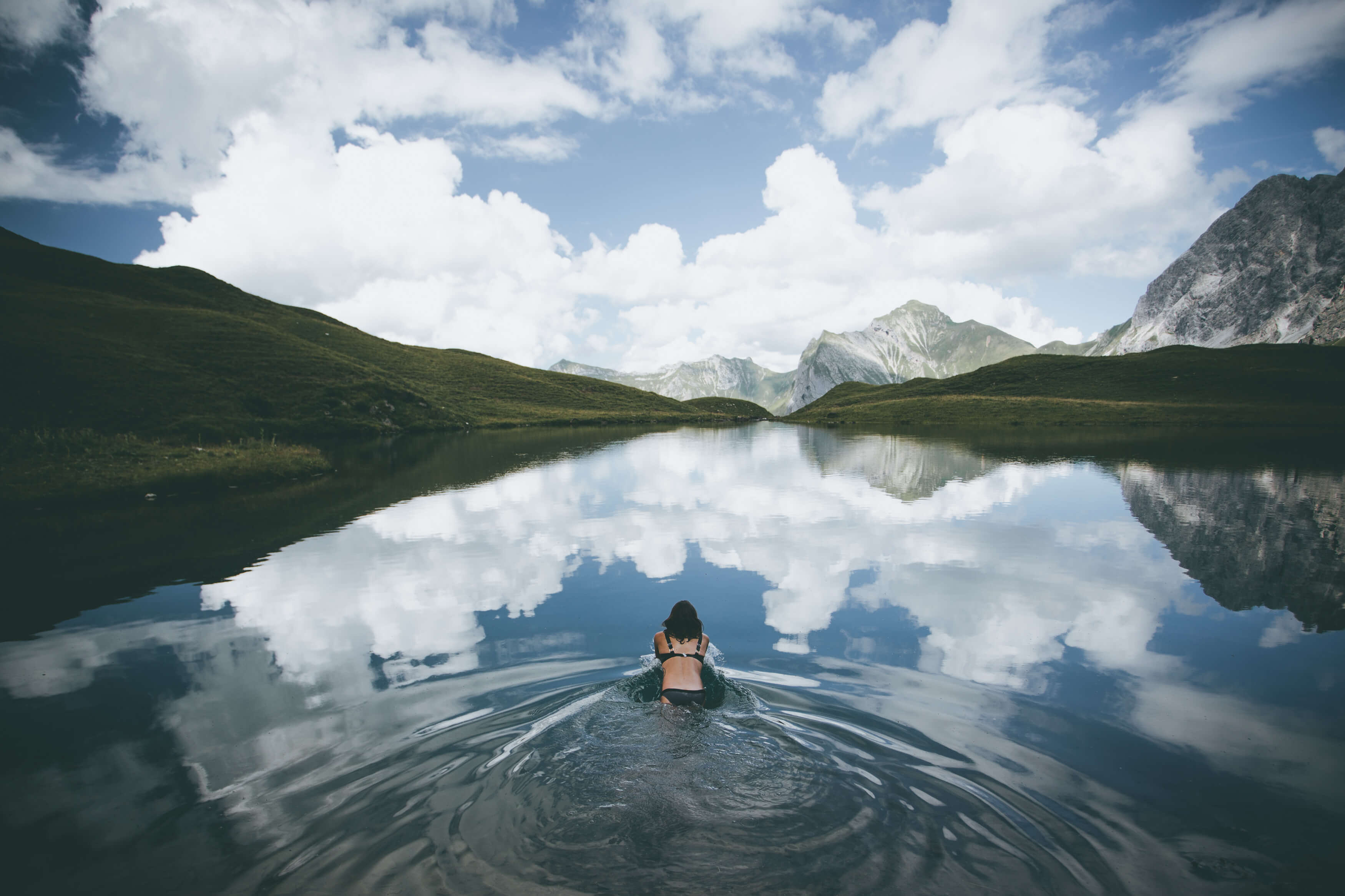 Schwimmerin von hinten in einem stillen Bergsee, in dem sich Wolken und Gipfel spiegeln