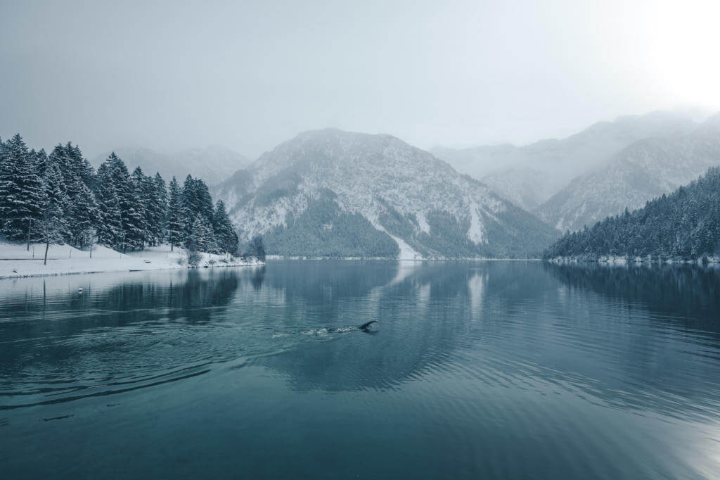 Weite Winterlandschaft am Plansee, im stillen Wasser krault eine einzelne Person vor verschneiten Bergen