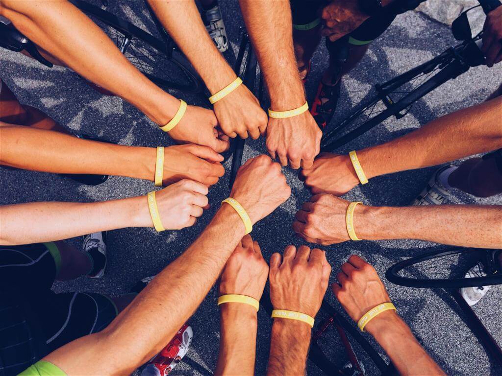 Viele Arme mit gelben Armbändern bilden über Asphalt einen Kreis, die Fäuste treffen sich in der Mitte