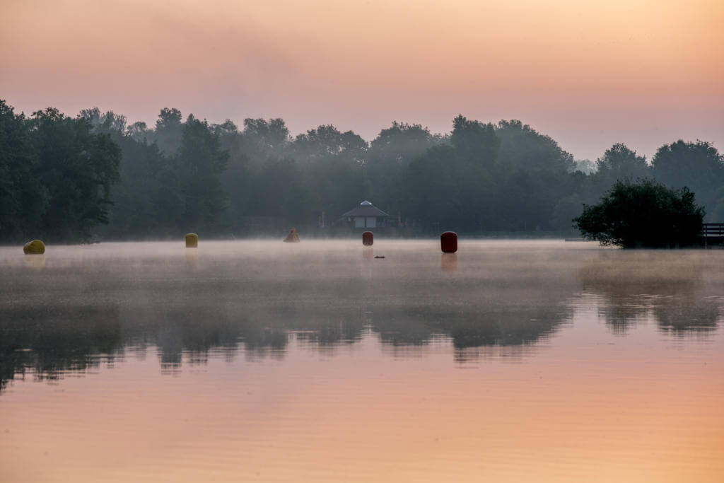Nebliger See im rosafarbenen Morgenlicht mit Bojen im Wasser und einem kleinen Pavillon am Ufer