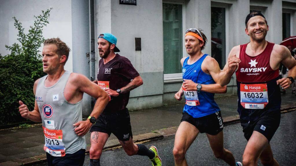 Vier Läufer mit Haspa-Startnummern beim Halbmarathon in Hamburg, rechts lacht ein Läufer im roten Trikot