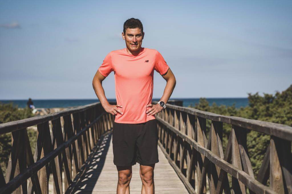 Triathlet Fabian Günther im lachsfarbenen Shirt steht mit den Händen in den Hüften auf einem Holzsteg am Meer