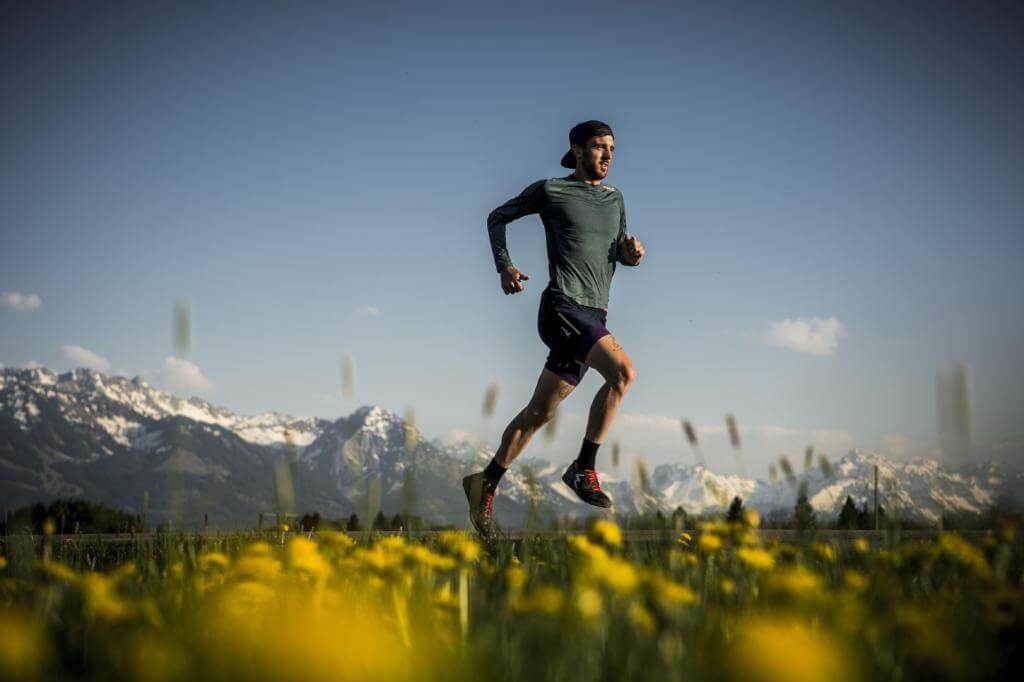 Läufer in dunkelgrünem Shirt im Laufschritt über eine gelb blühende Wiese vor schneebedeckten Bergen