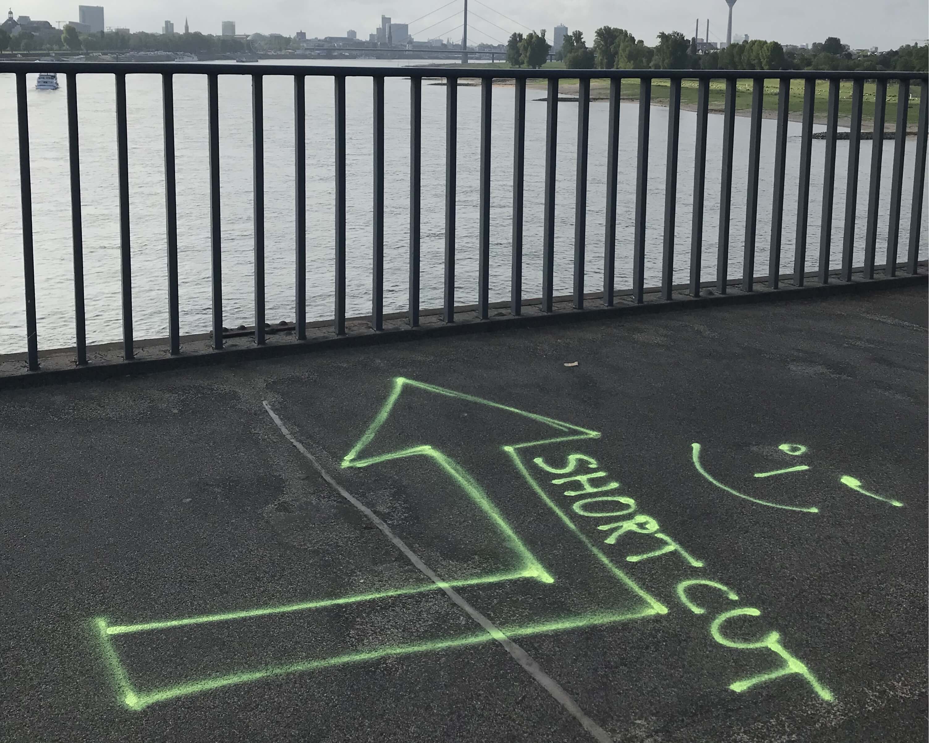 Mit neongelber Farbe auf den Asphalt gesprühter Pfeil mit der Aufschrift Short Cut, dahinter der Rhein in Düsseldorf