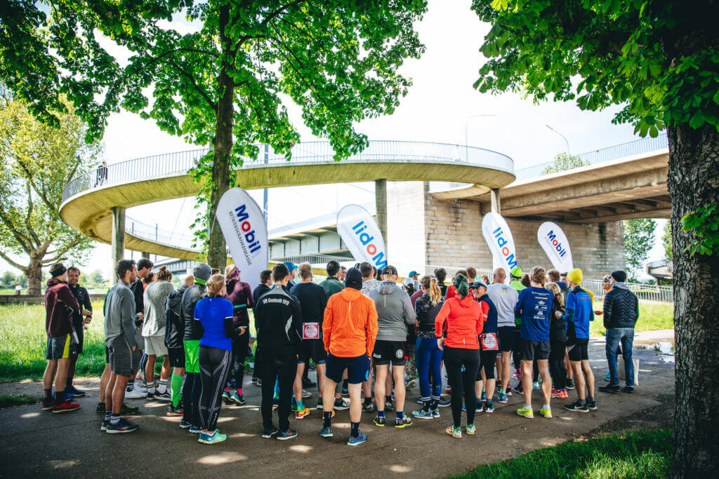 Teilnehmer versammeln sich zur Begrüßung vor Beachflags unter einer Brückenrampe, Wings for Life App Run Düsseldorf