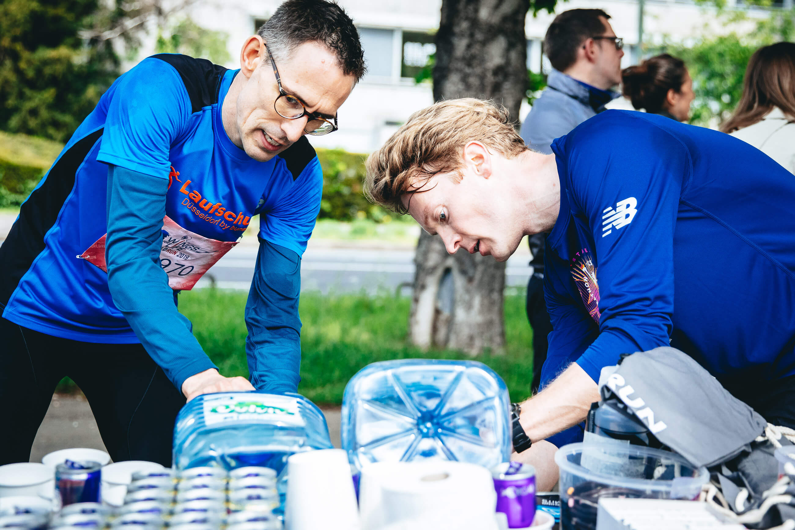 Zwei Läufer an einem Verpflegungstisch mit Wasserkanistern, Bechern und Dosen, Wings for Life App Run Düsseldorf