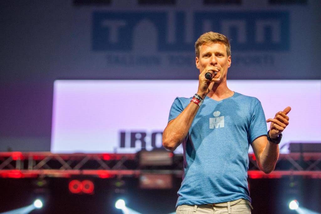 Till Schenk im blauen Ironman-Shirt spricht mit Mikrofon auf der Bühne beim Ironman Tallinn
