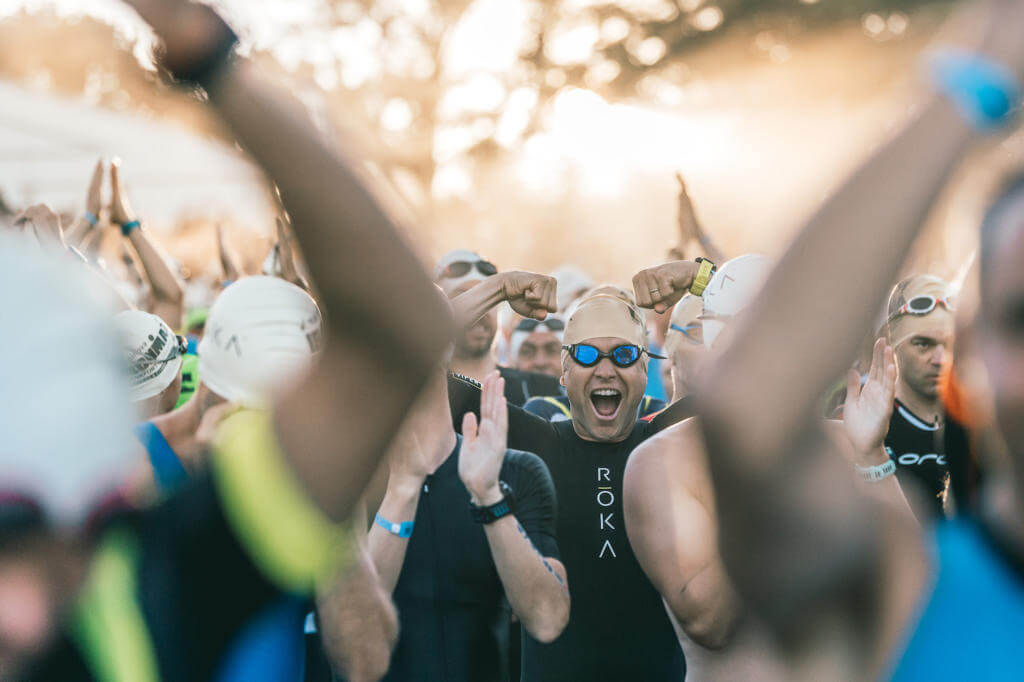 Jubelnde Athleten mit Badekappen und Schwimmbrillen im Gegenlicht vor dem Schwimmstart des Ironman Frankfurt 2019