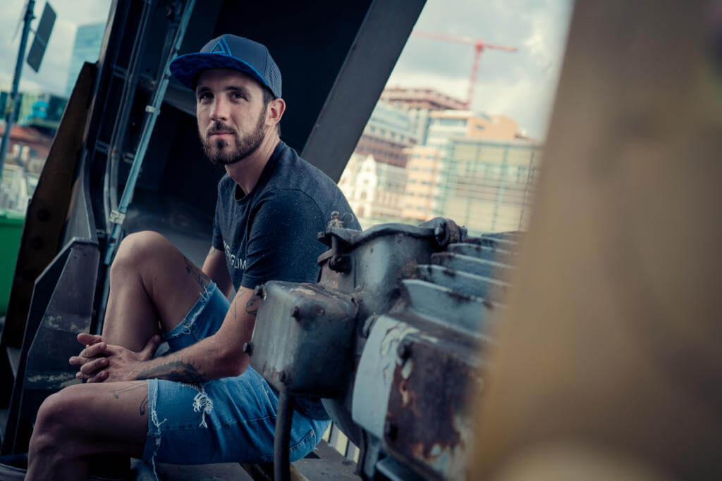 Niclas Bock mit der Pushing Limits Spirit Cap sitzt auf einem alten Kran am Hafen vor moderner Architektur