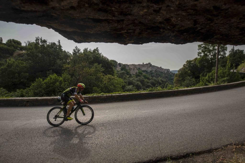 Triathlet auf der Radstrecke des Ironman France bei Nizza unter einem Felsüberhang, dahinter ein Bergdorf