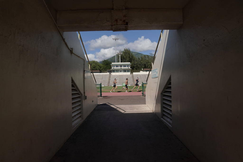 Blick durch einen dunklen Stadiontunnel auf drei Läufer beim Bahntraining auf Maui, dahinter Tribüne und Berge