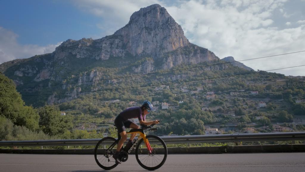 Radfahrerin auf einem gelb-roten Triathlonrad vor einem Felsgipfel an der Radstrecke der Ironman 70.3 WM in Nizza