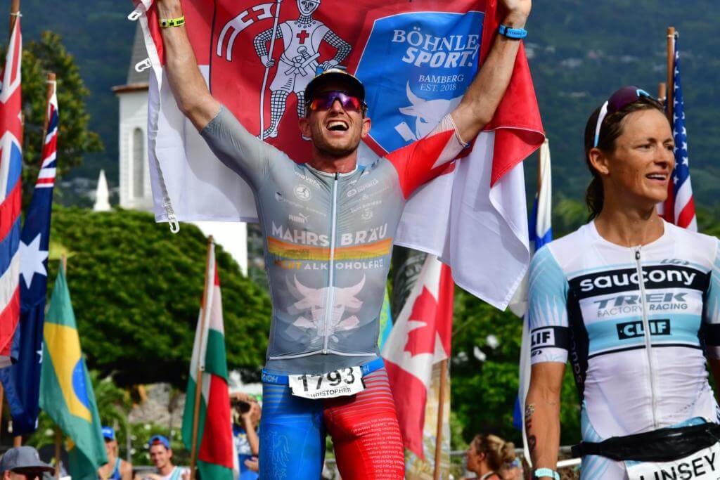 Christopher Dels jubelt mit einer Fahne über dem Kopf im Ziel des Ironman Hawaii vor Nationalflaggen