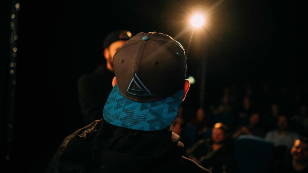 Person mit Pushing-Limits-Basecap von hinten vor Publikum in einem abgedunkelten Saal, dahinter ein Scheinwerfer