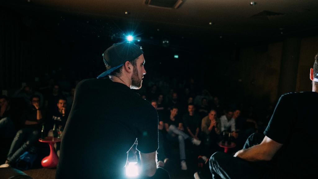 Moderator mit Basecap im Gegenlicht auf der Bühne, vor ihm das Publikum im Kinosaal bei der Get Together-Tour