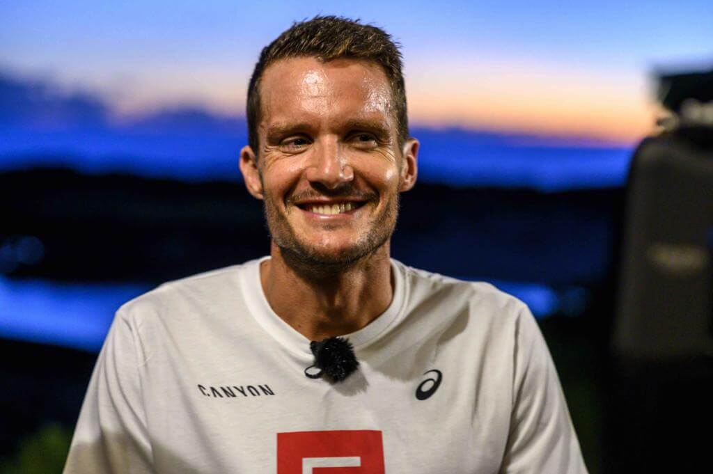 Jan Frodeno lächelt im weißen Canyon-Shirt mit Ansteckmikrofon vor dem Abendhimmel