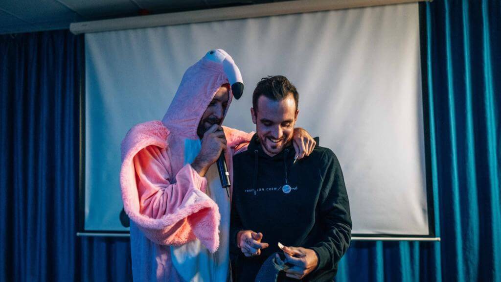 Mann im rosa Flamingo-Kostüm mit Mikrofon legt auf einer Bühne den Arm um einen lachenden Mann im schwarzen Hoodie