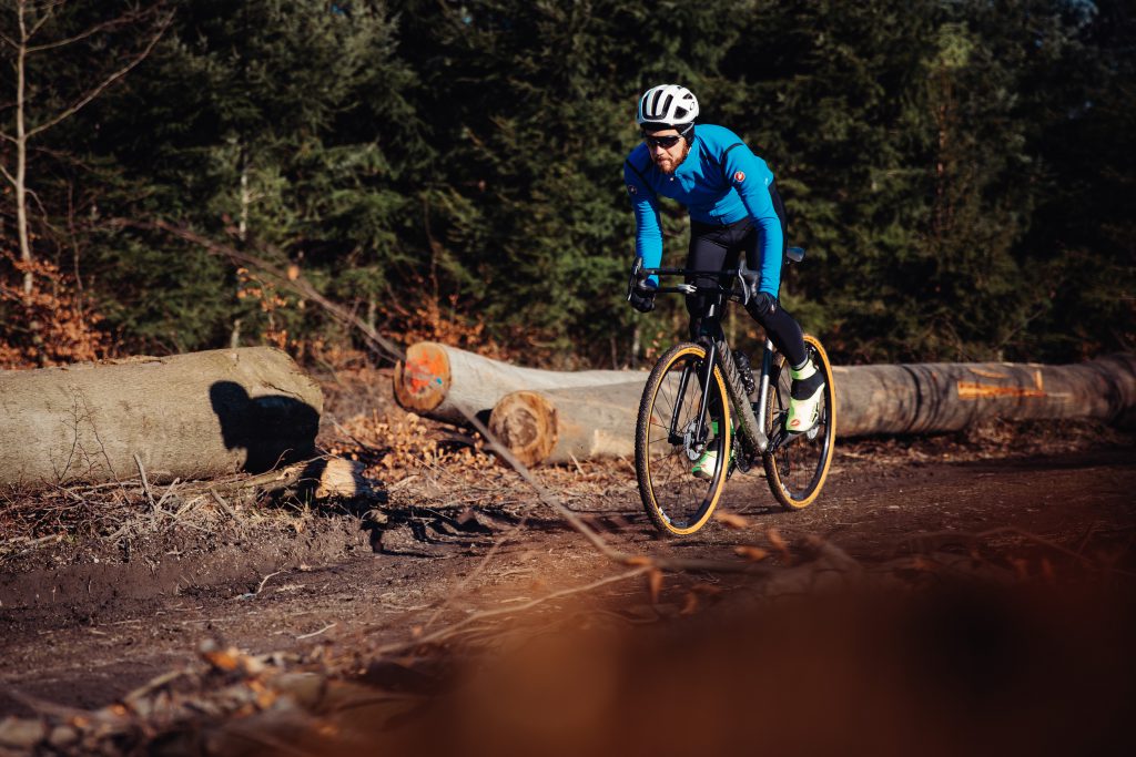 Radfahrer im blauen Trikot fährt mit dem Gravel-Bike auf einem Waldweg an gefällten Baumstämmen vorbei