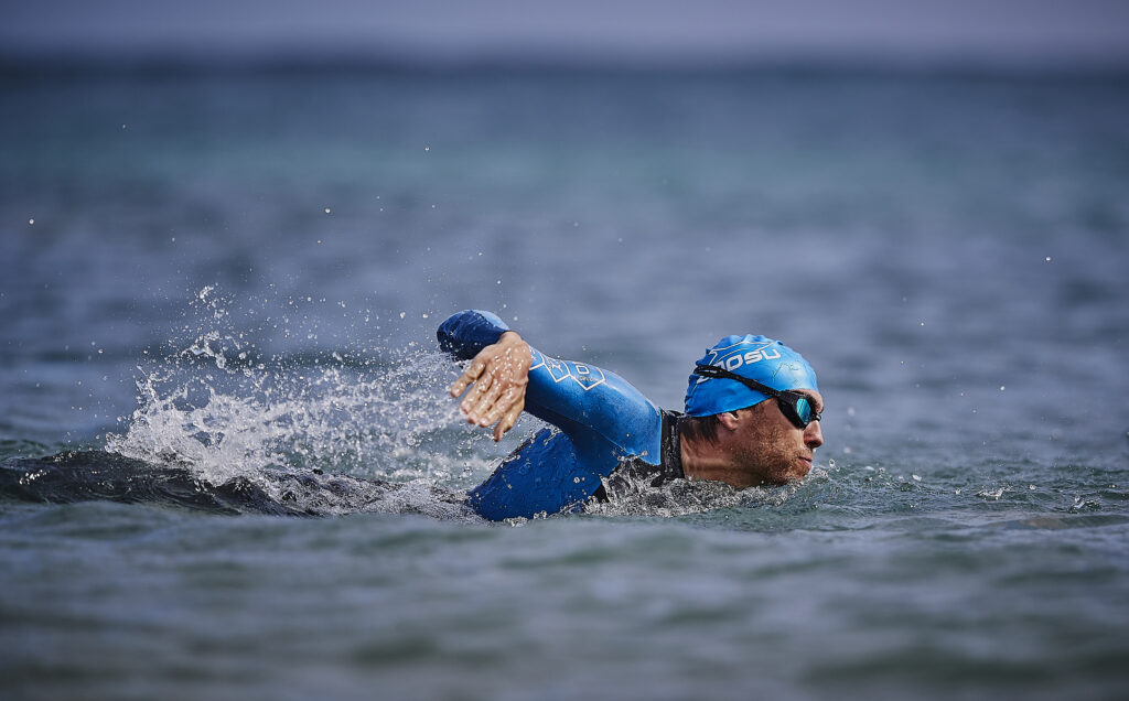 Schwimmer im blauen Neoprenanzug und blauer Zaosu-Badekappe krault im offenen Gewässer
