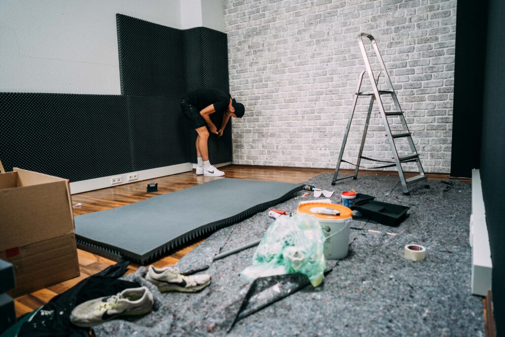 Umbau im neuen Studio mit Akustikschaumstoff, Farbeimer, Leiter und Malervlies, hinten ein Mann an der Wand