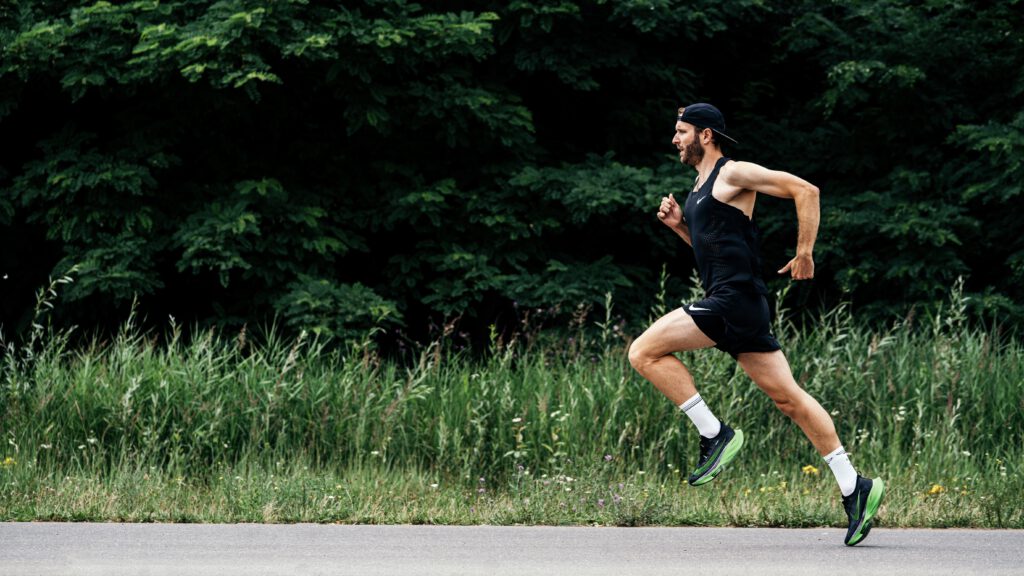 Läufer in schwarzem Tanktop und neongrünen Nike Alphafly beim schnellen Lauf auf einer Straße vor Bäumen