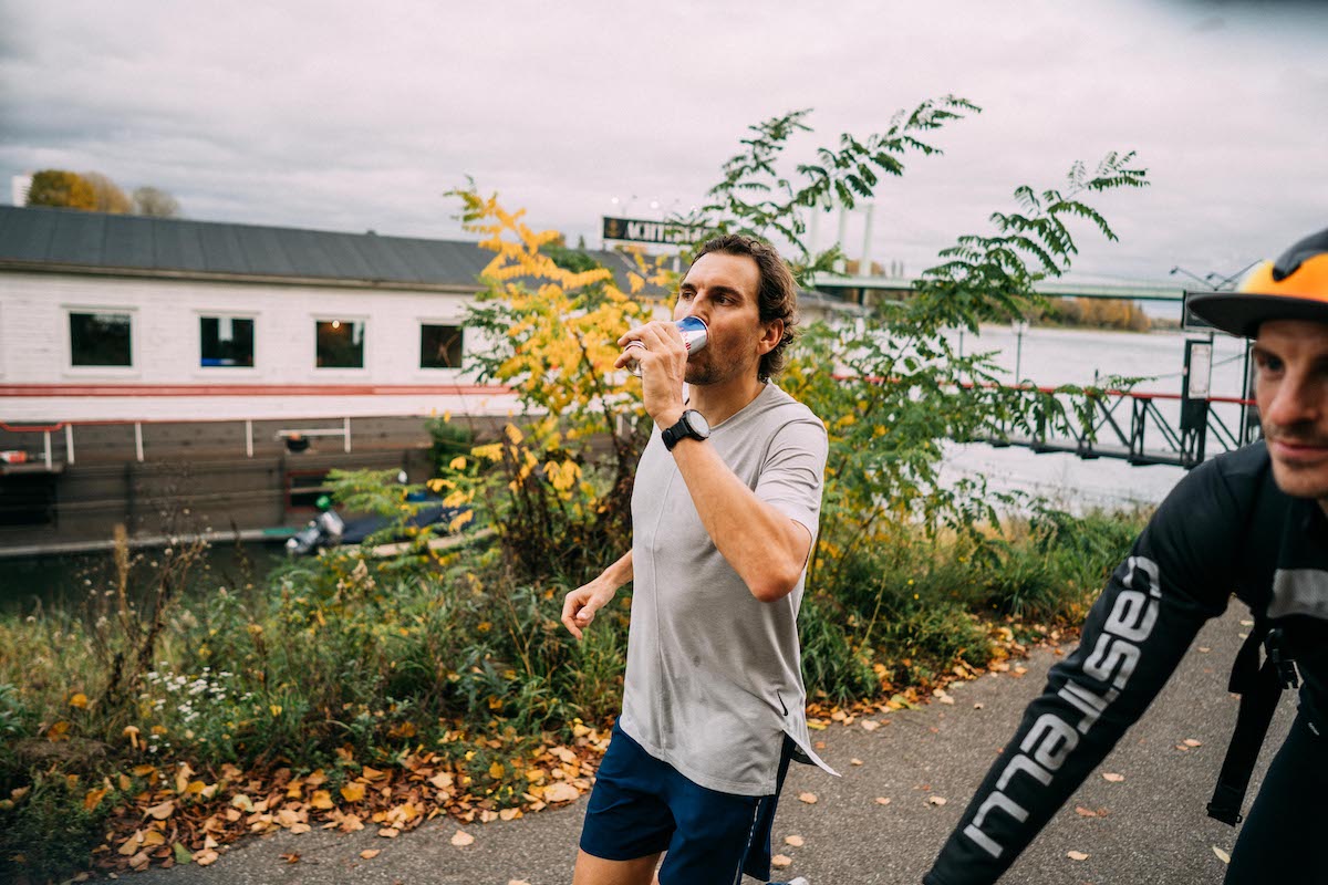Läufer trinkt beim Ultralauf am Rhein aus einer Red-Bull-Dose, rechts ein Radbegleiter
