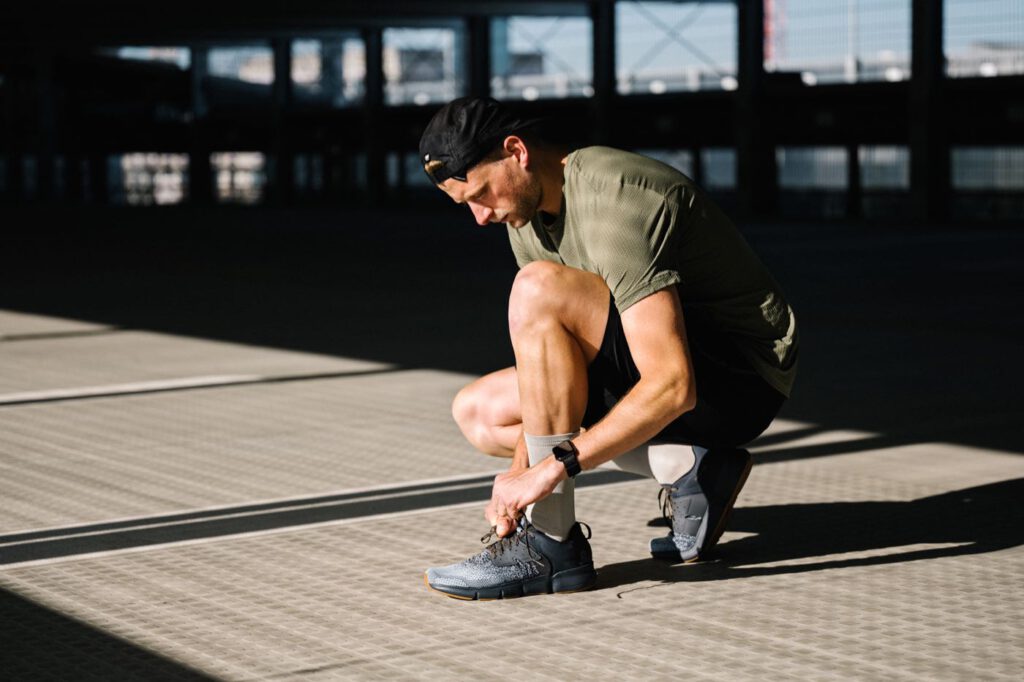 Läufer in olivgrünem Shirt und kurzer Hose von Fe226 bindet sich im Sonnenlicht die Laufschuhe zu