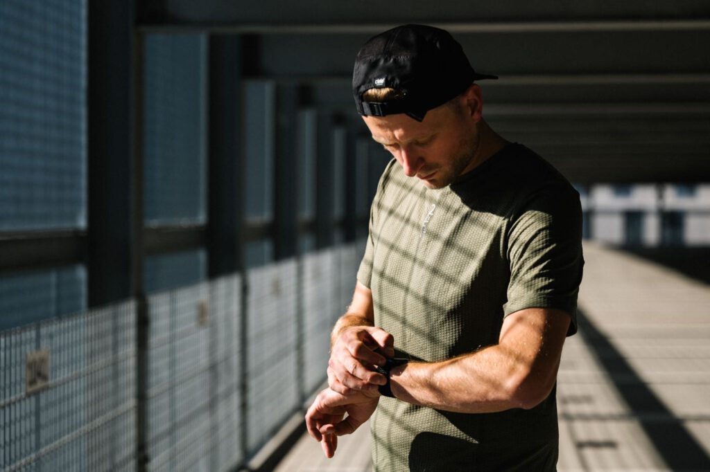 Läufer mit Kappe im olivgrünen TEM Running T-Shirt von Fe226 blickt auf einem überdachten Steg auf seine Sportuhr