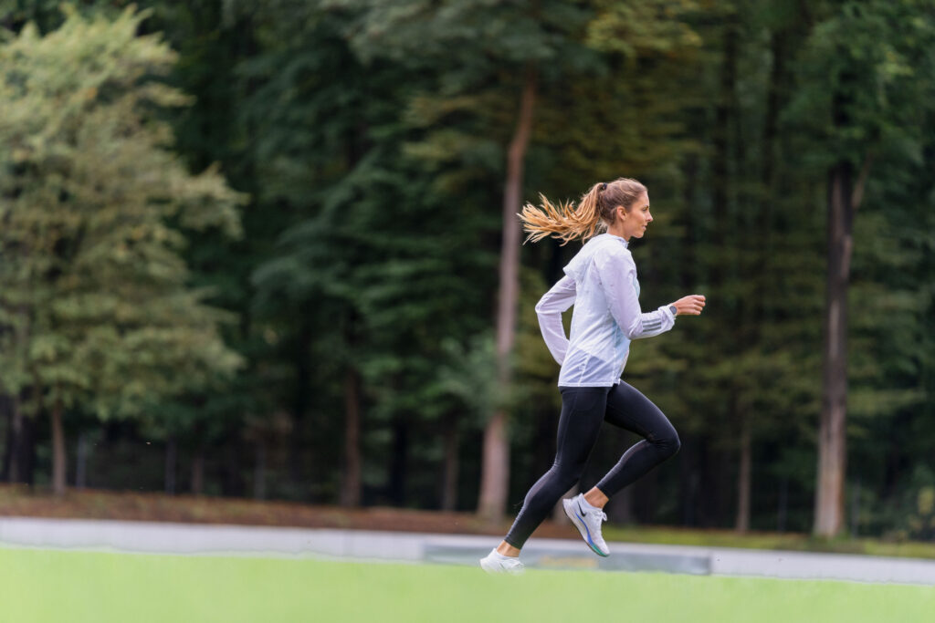 Läuferin in heller Jacke, schwarzer Tight und Laufschuhen vor einem Waldrand, Laufausrüstung für den ersten Triathlon