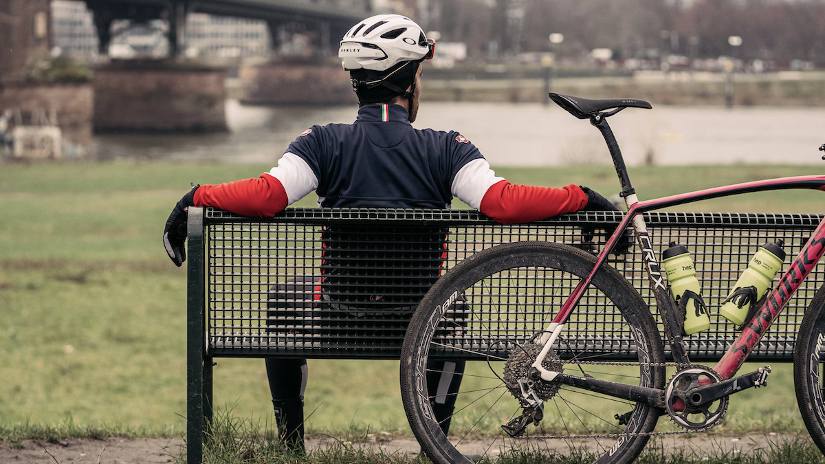Ein Radfahrer sitzt auf einer Bank an der sein Rad gelehnt ist und schaut auf einen Fluss