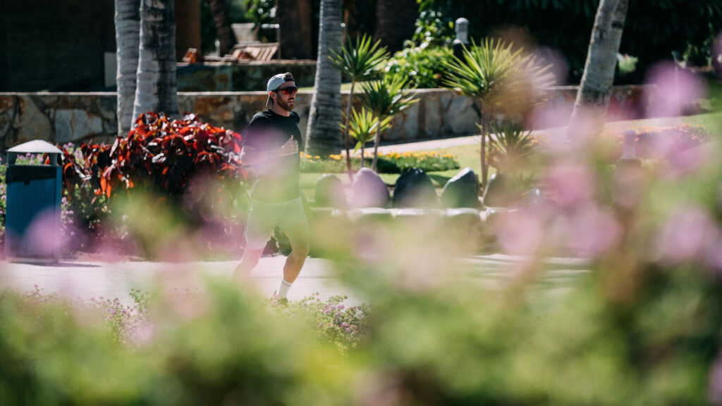 Läufer mit Cap und Sonnenbrille joggt durch eine Gartenanlage mit Palmen, vorne unscharfe rosa Blüten