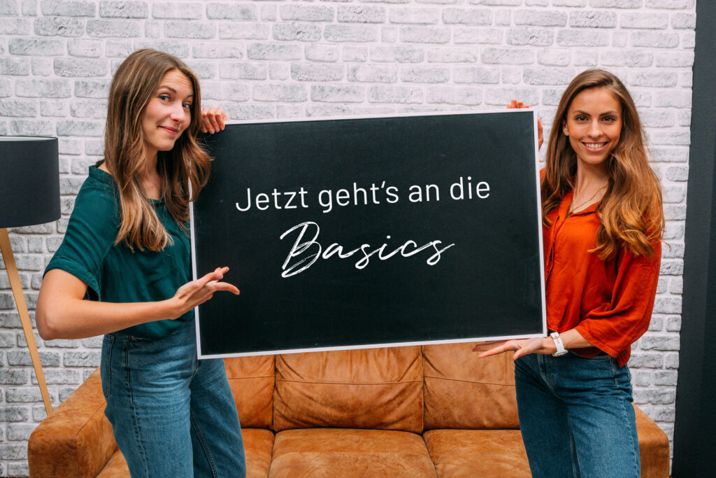 Zwei lächelnde Frauen halten vor einem Ledersofa eine Tafel mit der Aufschrift Jetzt geht's an die Basics